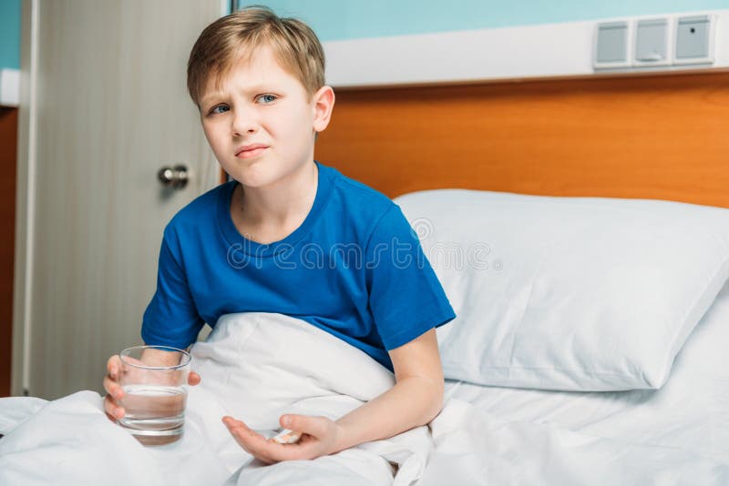 Retrato do menino insatisfeito que guarda o vidro da água e das medicinas na cama de hospital