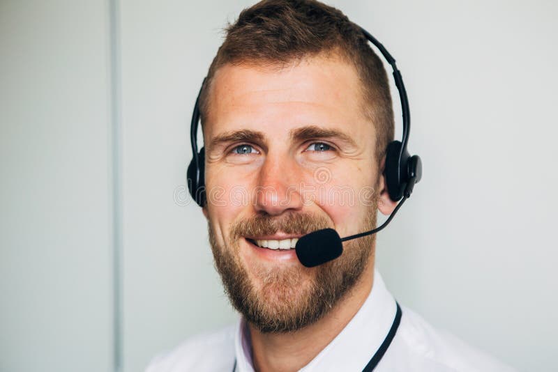 Retrato do operador masculino novo considerável nos auriculares que olham a câmera e que sorriem ao estar contra o branco imagem de stock royalty free Retrato do operador masculino novo considerável nos auriculares que olham a câmera e que sorriem ao estar contra o branco imagem de stock royalty free