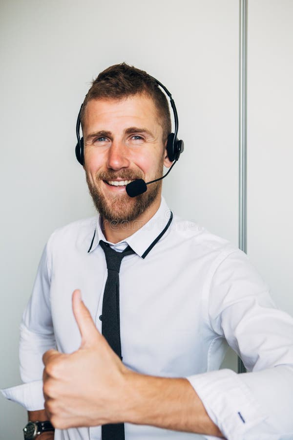 Retrato do operador masculino novo considerável nos auriculares que olham a câmera e que sorriem ao estar contra o branco fotografia de stock Retrato do operador masculino novo considerável nos auriculares que olham a câmera e que sorriem ao estar contra o branco fotografia de stock