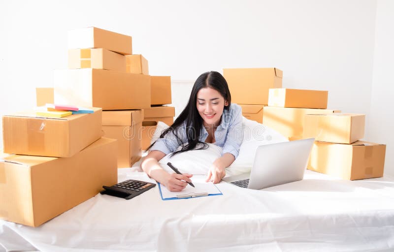 Retrato do portátil de trabalho da mulher asiática nova com o pacote postal na cama em casa, vendendo ideias em linha conceito e fotografia de stock royalty free Retrato do portátil de trabalho da mulher asiática nova com o pacote postal na cama em casa, vendendo ideias em linha conceito e fotografia de stock royalty free