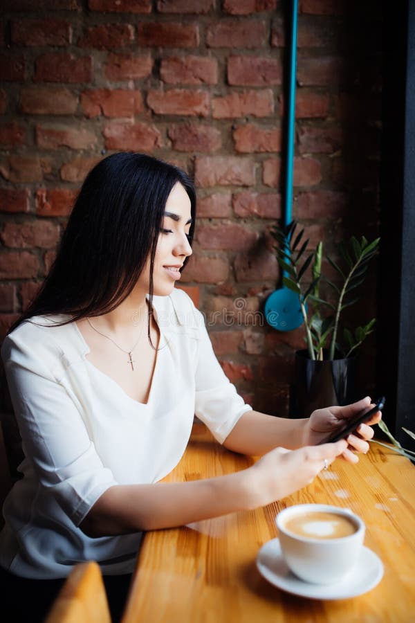 Retrato do telefone celular do uso da jovem mulher ao sentar-se na cafetaria confortável durante a ruptura de trabalho, leitura f fotografia de stock Retrato do telefone celular do uso da jovem mulher ao sentar-se na cafetaria confortável durante a ruptura de trabalho, leitura f fotografia de stock
