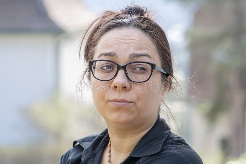 Retrato emocional de una disgustada ama de casa de 40 años con gafas en un fondo borroso de un paisaje suburbano.