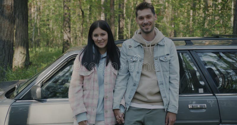 Retrato en cámara lenta de parejas de estudiantes felices parados al lado de un auto moderno sonriendo al aire libre en el bosque