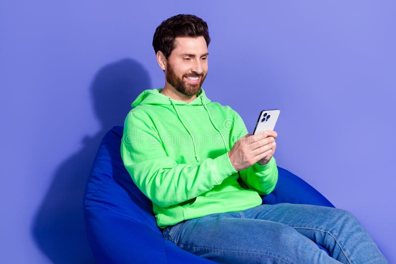 Retrato fotográfico de un hombre apuesto y maduro que usa una capucha verde brillante tumbada en un puf y habla con un smartphone imagen de archivo