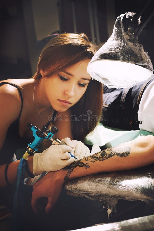 Retrato hermoso del artista del tatuaje de la chica joven durante tatuaje de la creación en una mano del ` s del hombre foto de archivo libre de regalías Retrato hermoso del artista del tatuaje de la chica joven durante tatuaje de la creación en una mano del ` s del hombre foto de archivo libre de regalías