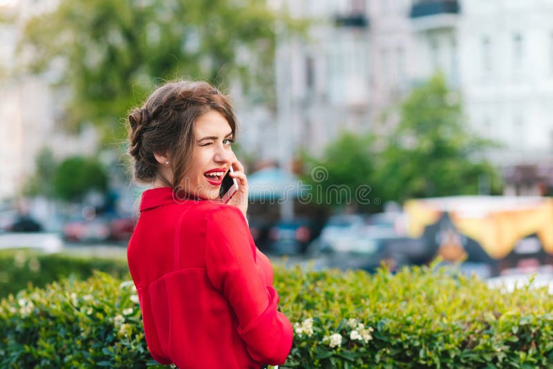 Retrato horizontal da posição bonita da menina no parque Veste a blusa vermelha e o penteado agradável Está falando no telefone imagem de stock royalty free