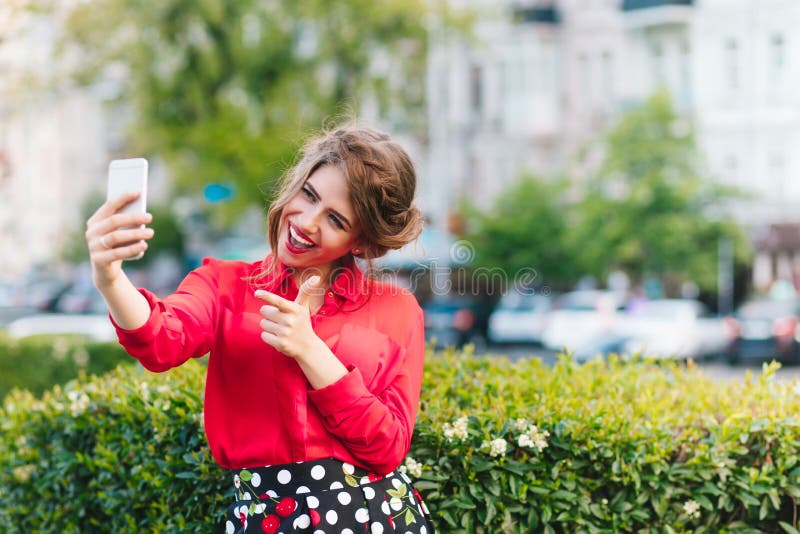 Retrato horizontal da posição bonita da menina no parque Veste a blusa vermelha e o penteado agradável Está fazendo um selfie fotografia de stock
