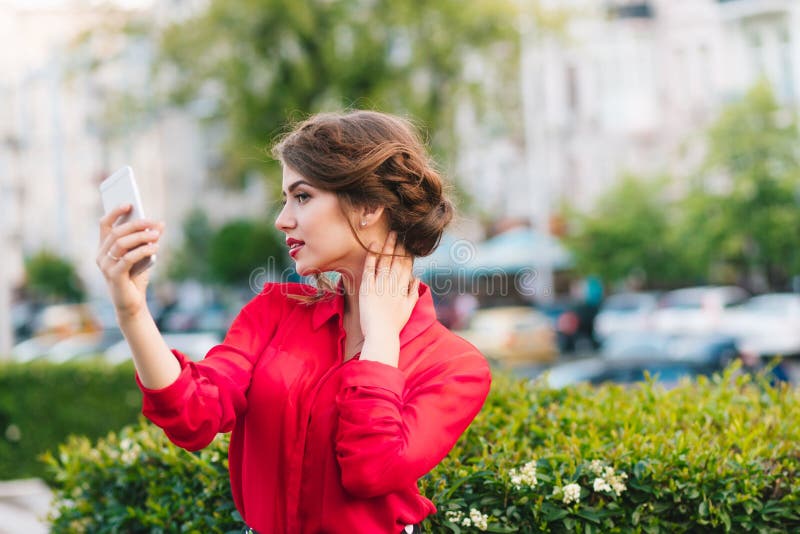 Retrato horizontal da posição bonita da menina no parque Veste a blusa vermelha e o penteado agradável Está usando um telefone de fotos de stock