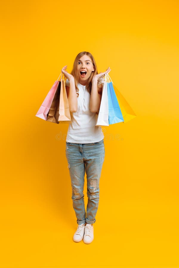 Retrato integral de una mujer chocada joven, feliz después de hacer compras con los bolsos multicolores, en un fondo amarillo imagen de archivo libre de regalías Retrato integral de una mujer chocada joven, feliz después de hacer compras con los bolsos multicolores, en un fondo amarillo imagen de archivo libre de regalías