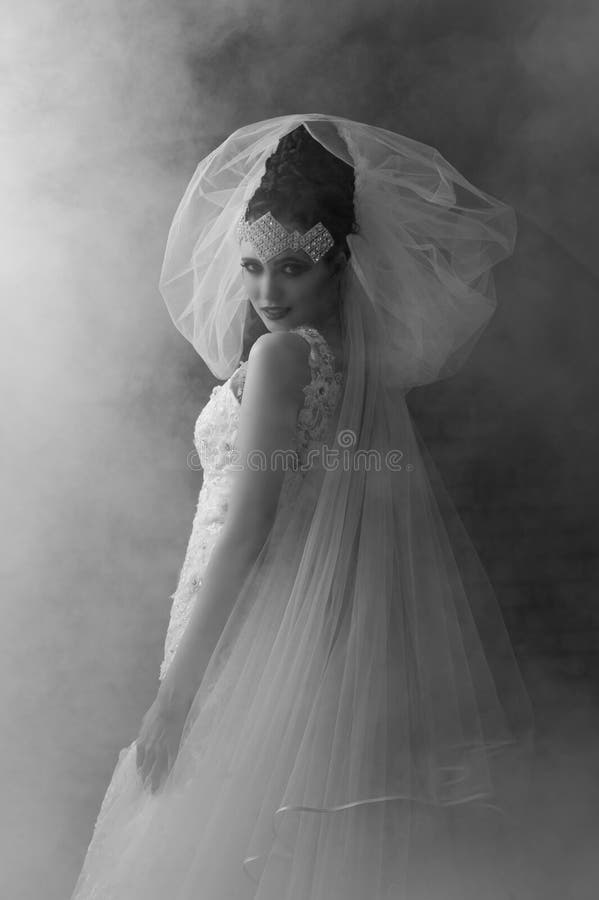 Portrait in monochrome of a beautiful and dramatic bride wearing a wedding dress with sparkling diamonds and embroidery, dramatic makeup, a bunched tulle veil with her dark hair in a dramatic braided hairstyle with a diamond crown headdress, standing looking back over her shoulder with a smile while a mysterious smoky mist surrounds her. Portrait in monochrome of a beautiful and dramatic bride wearing a wedding dress with sparkling diamonds and embroidery, dramatic makeup, a bunched tulle veil with her dark hair in a dramatic braided hairstyle with a diamond crown headdress, standing looking back over her shoulder with a smile while a mysterious smoky mist surrounds her.