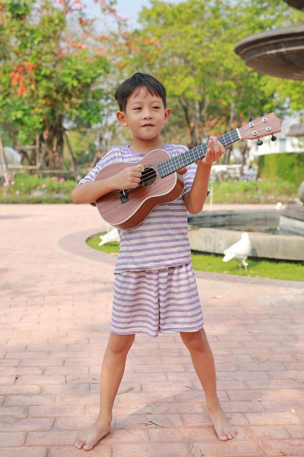 Retrato niño asiático divirtiéndose jugando al clásico ukulele mientras está parado en el jardín de verano imagen de archivo libre de regalías
