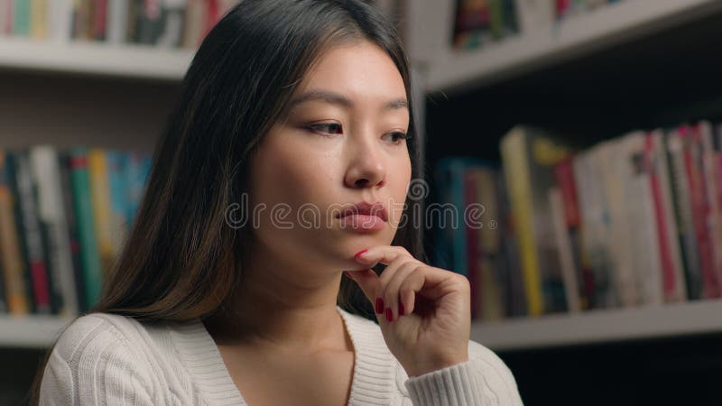 Retrato pensativo e pensivo da mulher empresária asiática estudante feminina na biblioteca perto do pôster da estante de livraria