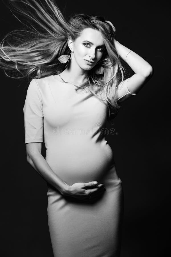 Black and white portrait of beautiful mom embracing stomach, smiling, looking away, over shoulder. Young happy mother posing at studio, against dark background. Pregnant fashion. Black and white portrait of beautiful mom embracing stomach, smiling, looking away, over shoulder. Young happy mother posing at studio, against dark background. Pregnant fashion.