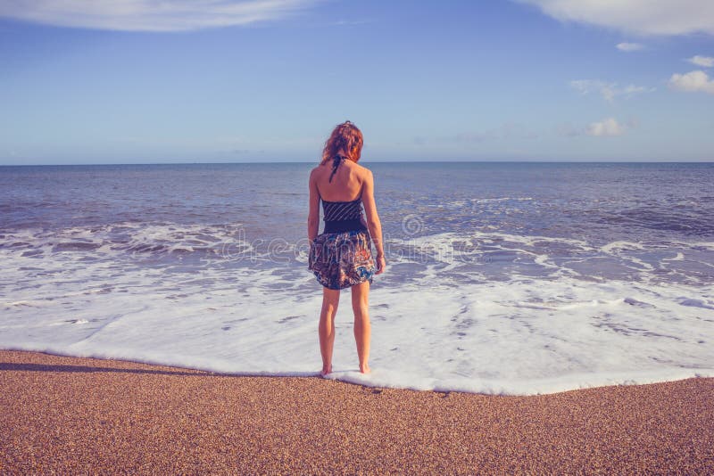 Retrovisione della giovane donna che sta sulla spiaggia che esamina mare immagini stock Retrovisione della giovane donna che sta sulla spiaggia che esamina mare immagini stock