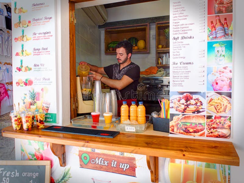 RHODES Ö, LINDOS, GREKLAND, JUNI, 25, 2015: Den grekiska bartendermannen i litet gatakafé med nya fruktsafter, mjölkar och frukts arkivfoton RHODES Ö, LINDOS, GREKLAND, JUNI, 25, 2015: Den grekiska bartendermannen i litet gatakafé med nya fruktsafter, mjölkar och frukts arkivfoton