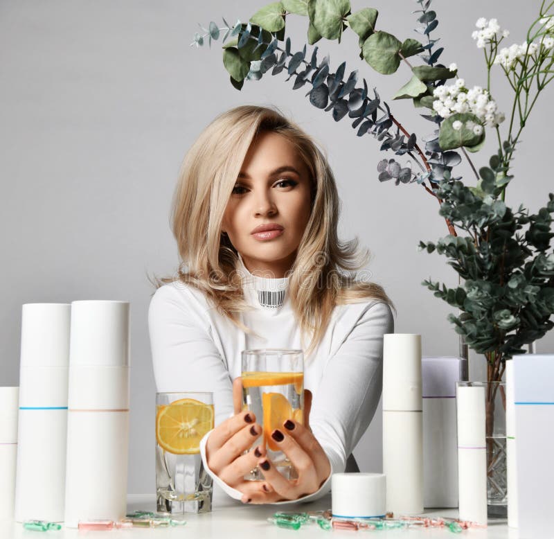 Rich blonde woman in white pullover sits at table with jars and tubes of cosmetic means, holding glass of lemon water