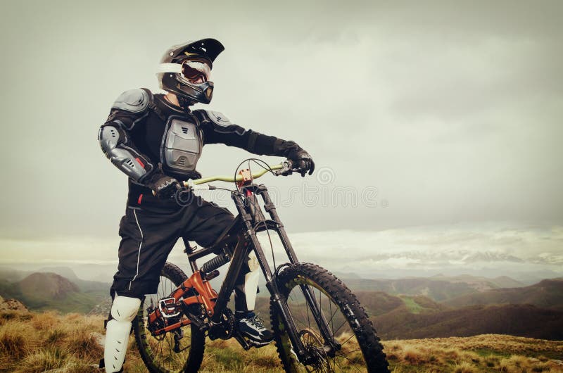 The rider in the full-face helmet and full protective equipment on the mtb bike stands on a rock against the background