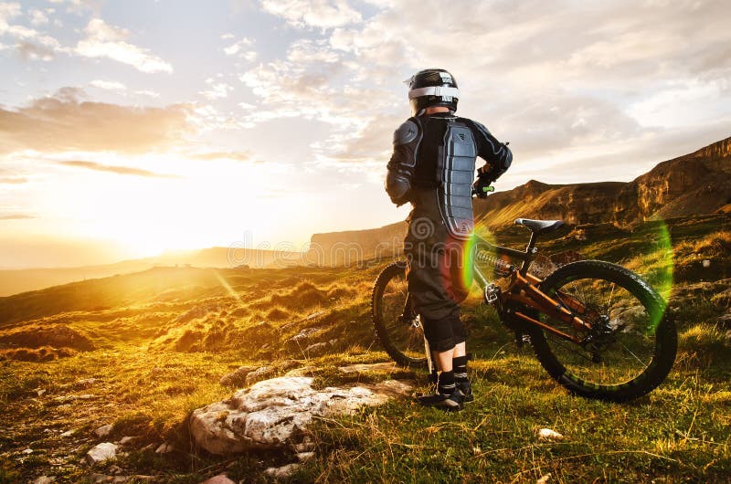 The rider in full protective equipment on the mtb bike is riding toward the sunset in the rays of the sunset sun against
