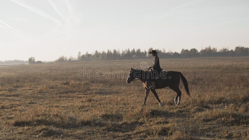 Ridhäst vid solnedgång i fält. cowgirl i hatt vid brun häst