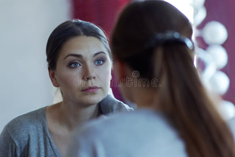 Riflessione di giovane donna caucasica attraente che esamina uno specchio Uso degli occhi azzurri casuali e bei, sguardo serio
