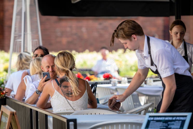 RIGA LETTLAND - JULI 31, 2018: Utomhus- café på tabellen sitter en kvinna med en stor tatuering på hans baksida bak parman tjäna royaltyfri fotografi RIGA LETTLAND - JULI 31, 2018: Utomhus- café på tabellen sitter en kvinna med en stor tatuering på hans baksida bak parman tjäna royaltyfri fotografi