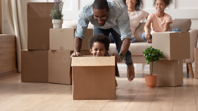 Happy african american little boy sitting in cardboard box while smiling barefoot father pushing it, moving, playing with small son in new living room, unpacking, relocation, new house purchase. Happy african american little boy sitting in cardboard box while smiling barefoot father pushing it, moving, playing with small son in new living room, unpacking, relocation, new house purchase.