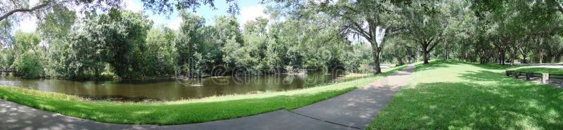 A panoramic photo of a river next to a sidewalk and surrounded by trees. A panoramic photo of a river next to a sidewalk and surrounded by trees.