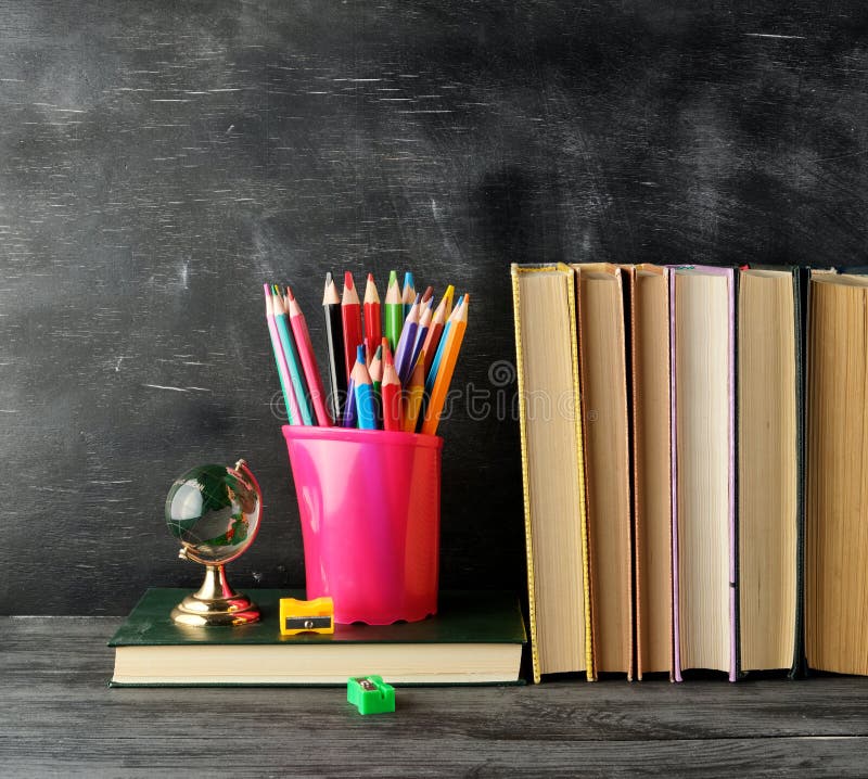 stack of books and a blue stationery glass with multi-colored wooden pencils,  on the background of an empty black chalk board, back to school concept. stack of books and a blue stationery glass with multi-colored wooden pencils,  on the background of an empty black chalk board, back to school concept