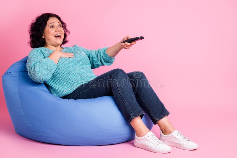 Ritratto completo della donna interna seduta in poltrona e tenuta il telecomando della tv su sfondo pastello immagine stock libera da diritti