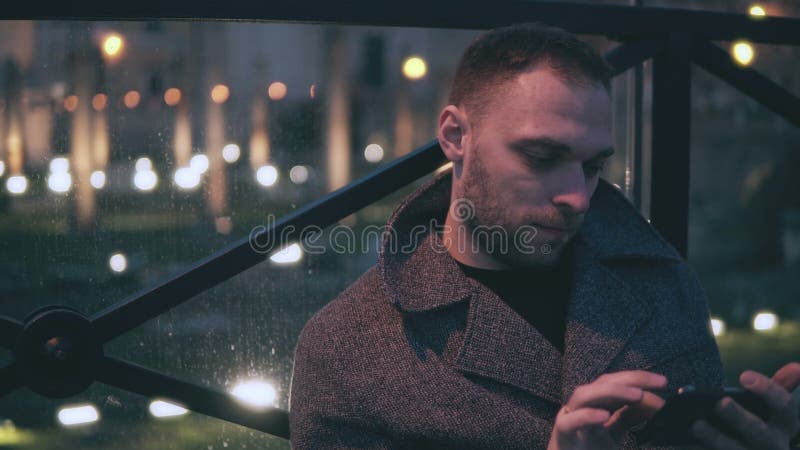 Ritratto del giovane attraente che si siede nella città di sera e nello smartphone di uso Il maschio bello passa in rassegna Inte immagini stock libere da diritti Ritratto del giovane attraente che si siede nella città di sera e nello smartphone di uso Il maschio bello passa in rassegna Inte immagini stock libere da diritti