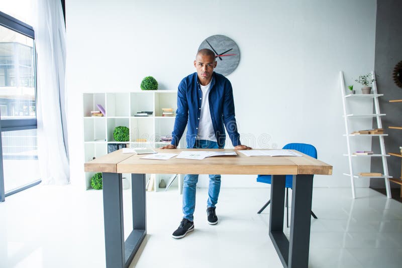 Ritratto del giovane sicuro che sta al suo scrittorio con il computer portatile Uomo d'affari africano che lavora nell'ufficio mo immagine stock Ritratto del giovane sicuro che sta al suo scrittorio con il computer portatile Uomo d'affari africano che lavora nell'ufficio mo immagine stock