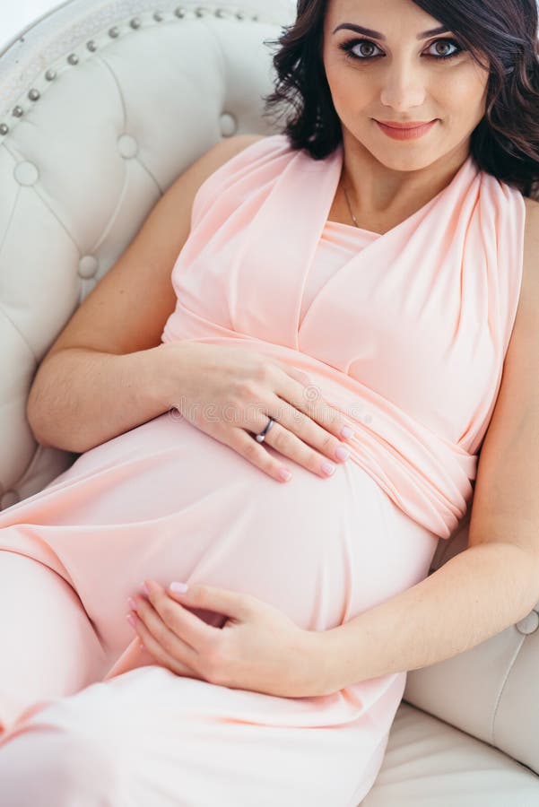 Ritratto del primo piano di bella donna incinta con il sorriso grazioso in vestito rosa che tocca la sua pancia mentre trovandosi immagine stock Ritratto del primo piano di bella donna incinta con il sorriso grazioso in vestito rosa che tocca la sua pancia mentre trovandosi immagine stock