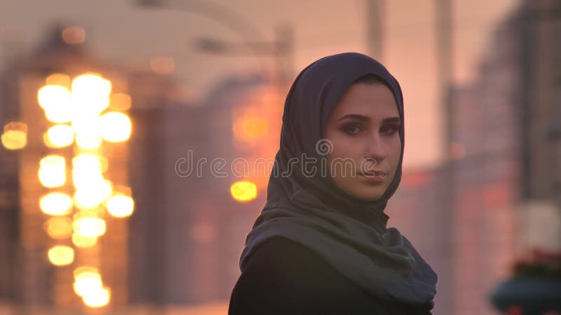 Ritratto del primo piano di giovane femmina graziosa nel hijab che gira e che esamina macchina fotografica con le costruzioni bri immagini stock Ritratto del primo piano di giovane femmina graziosa nel hijab che gira e che esamina macchina fotografica con le costruzioni bri immagini stock