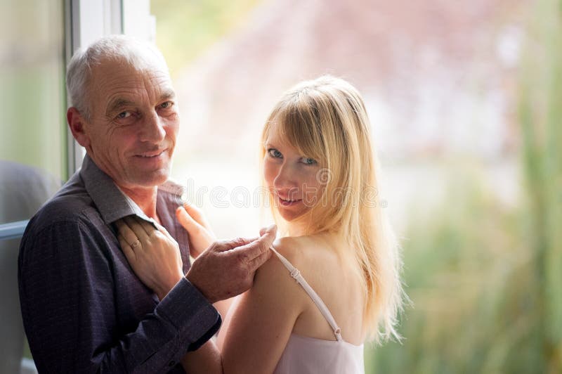Ritratto dell'uomo anziano che sta vicino alla finestra con la sua giovane moglie Bionda-dai capelli in vestito da short di estat fotografia stock Ritratto dell'uomo anziano che sta vicino alla finestra con la sua giovane moglie Bionda-dai capelli in vestito da short di estat fotografia stock