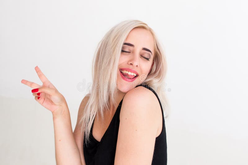 Portrait of cheerful woman with peace hand sign. Drunk playful blonde in black dress with closed eyes laughing and posing jocosely at camera, white background. Portrait of cheerful woman with peace hand sign. Drunk playful blonde in black dress with closed eyes laughing and posing jocosely at camera, white background