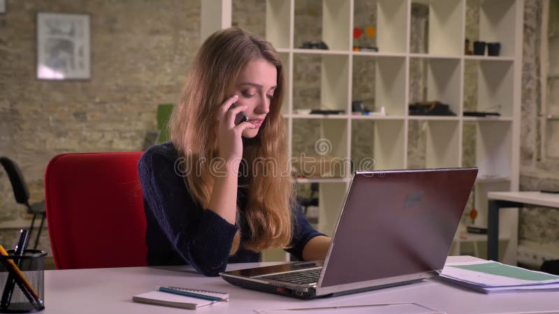 Ritratto della donna di affari caucasica bionda attenta davanti al computer portatile in ufficio che parla seriamente sul cellula