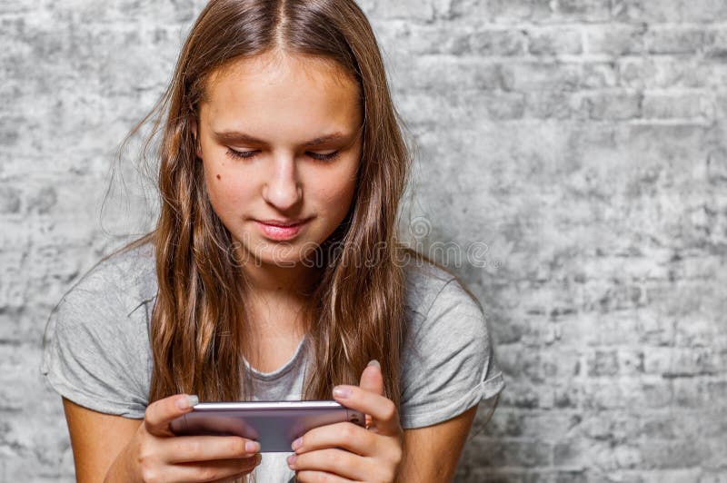 Ritratto della ragazza castana del giovane adolescente con capelli lunghi facendo uso del telefono cellulare sulla parete grigia fotografie stock Ritratto della ragazza castana del giovane adolescente con capelli lunghi facendo uso del telefono cellulare sulla parete grigia fotografie stock