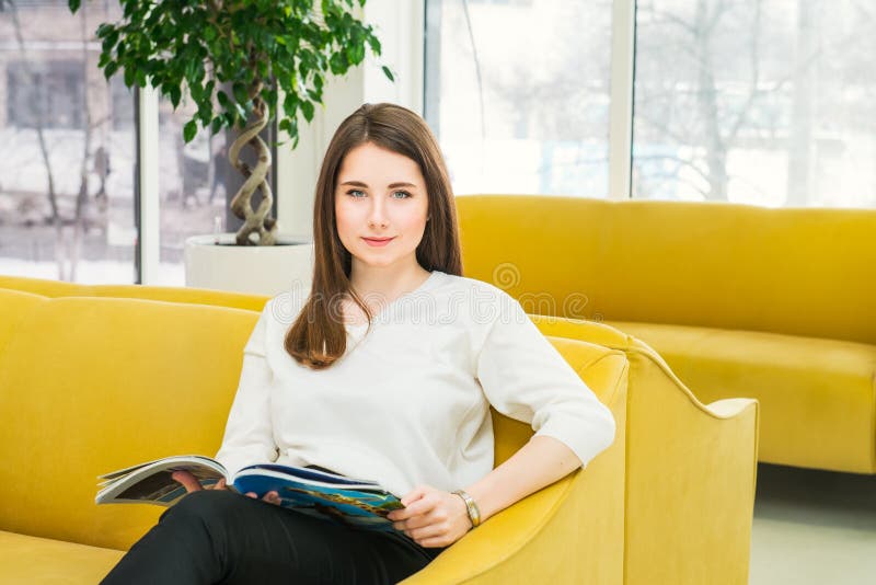 Ritratto della ragazza che esamina macchina fotografica, sedentesi sul sofà giallo luminoso in corridoio aspettante moderno e leg fotografia stock libera da diritti Ritratto della ragazza che esamina macchina fotografica, sedentesi sul sofà giallo luminoso in corridoio aspettante moderno e leg fotografia stock libera da diritti