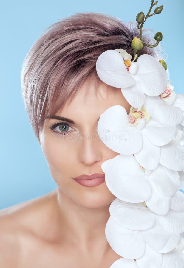 Ritratto di bella donna in un salone della stazione termale con l'orchidea bianca in sua mano su un fondo blu fotografia stock libera da diritti Ritratto di bella donna in un salone della stazione termale con l'orchidea bianca in sua mano su un fondo blu fotografia stock libera da diritti