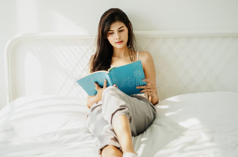 Ritratto di bella giovane donna asiatica che legge un libro in appartamento soleggiato e accogliente nella stanza bianca Rilassam immagine stock libera da diritti Ritratto di bella giovane donna asiatica che legge un libro in appartamento soleggiato e accogliente nella stanza bianca Rilassam immagine stock libera da diritti