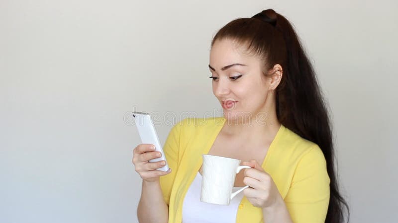 Ritratto di bella ragazza castana che beve una bevanda calda da una tazza e tiene un telefono cellulare in sua mano giovane