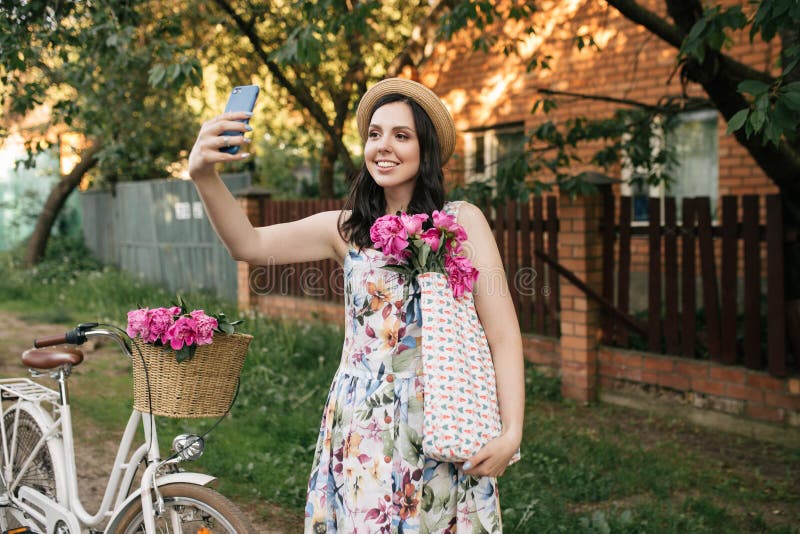Ritratto di bella ragazza felice con la bicicletta d'annata ed i fiori che fanno selfie Bici con il canestro pieno del rosa immagine stock Ritratto di bella ragazza felice con la bicicletta d'annata ed i fiori che fanno selfie Bici con il canestro pieno del rosa immagine stock