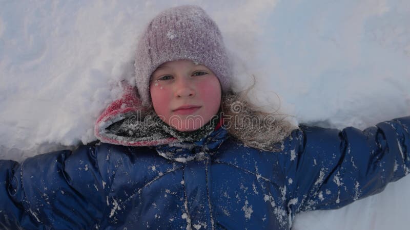Ritratto di chiusura di una bambina adorabile felice che sorride sdraiata sulla neve. Movimento lento stock footage