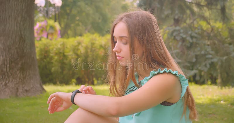 Ritratto di chiusura di una giovane e bella femmina con i capelli lunghi seduta sull'erba, con l'ausilio di un orologio elegante