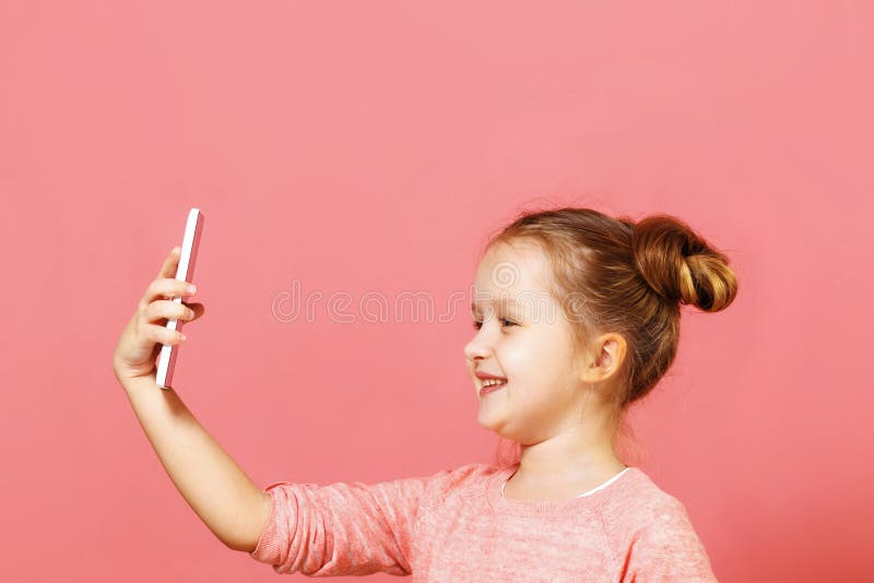 Ritratto di chiusura di una ragazzina carina con panini di capelli su uno sfondo rosa Il bambino tiene il telefono e si fa un sel immagini stock Ritratto di chiusura di una ragazzina carina con panini di capelli su uno sfondo rosa Il bambino tiene il telefono e si fa un sel immagini stock