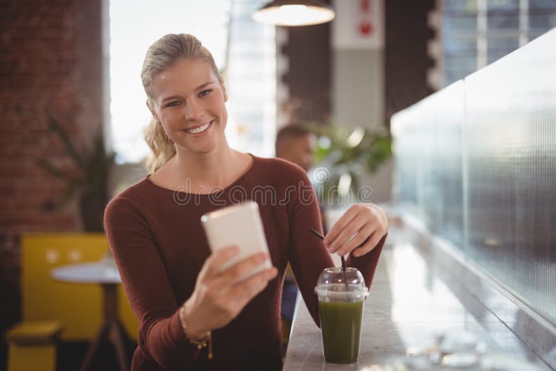 Ritratto di giovane donna bionda sorridente che per mezzo dello smartphone mentre sedendosi con la bevanda immagine stock Ritratto di giovane donna bionda sorridente che per mezzo dello smartphone mentre sedendosi con la bevanda immagine stock