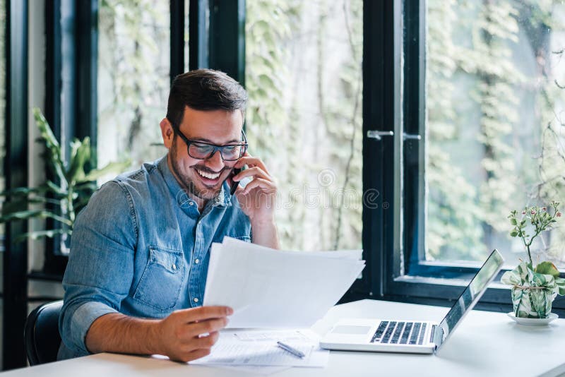 Ritratto di giovane imprenditore allegro sorridente in ufficio casuale che fa telefonata mentre lavorando con i grafici ed i graf