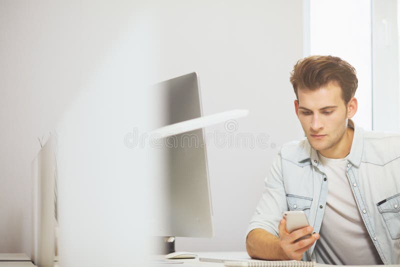 Ritratto di giovane uomo d'affari bello che si siede alla tavola dell'ufficio con il personal computer e che scrive sul telefono immagine stock Ritratto di giovane uomo d'affari bello che si siede alla tavola dell'ufficio con il personal computer e che scrive sul telefono immagine stock