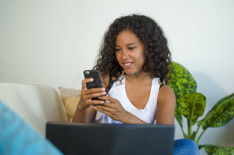 Lifestyle portrait of young happy and beautiful black african American woman using internet mobile phone while working on laptop computer at home living room sofa couch relaxed. Lifestyle portrait of young happy and beautiful black african American woman using internet mobile phone while working on laptop computer at home living room sofa couch relaxed