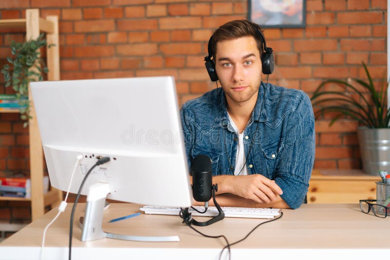 Ritratto di un bel blogger seducente, maschio, che indossa cuffie sedute sulla scrivania con microfono professionale e fotografia stock